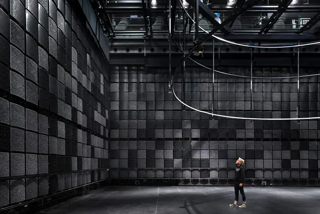 A white person standing in a black box studio looking at a wall of black sound proofing squares.