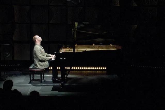 A man lifting his head towards the ceiling with eyes closed in a moment of reverence as he plays a grand piano on a dark stage in a spotlight. 