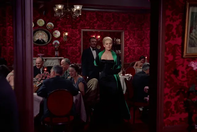 Kim Novak wearing a green 1950's cocktail dress entering a crowded restaurant with red damask wallpaper.