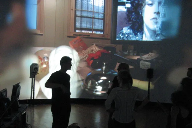 Three people chatting intently silhouetted against a large projection of a cluttered room. 