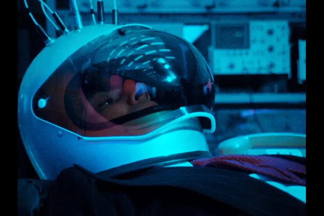 A man in a suit wearing an astronaut helmet reclined in room with various nobs and buttons washed in teal light. 