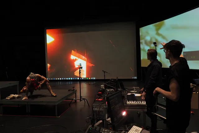 A performer in a squatting position on a black platform wearing a mask of sensors in front of two large screens projecting red and blue abstract scenes. Two people look on, one directing the scene with arm outstretched toward the screens. 
