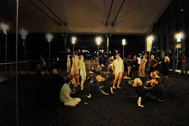 Dancers wearing white drapey unitards standing in contrapposto poses in the EMPAC lobby, light with warm lighting. A small crowd watches, seated on a black rug.  