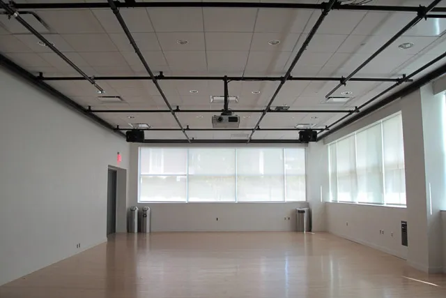 An empty studio with light wood floors, white walls, and black rigging on the ceiling flooded with natural light. 