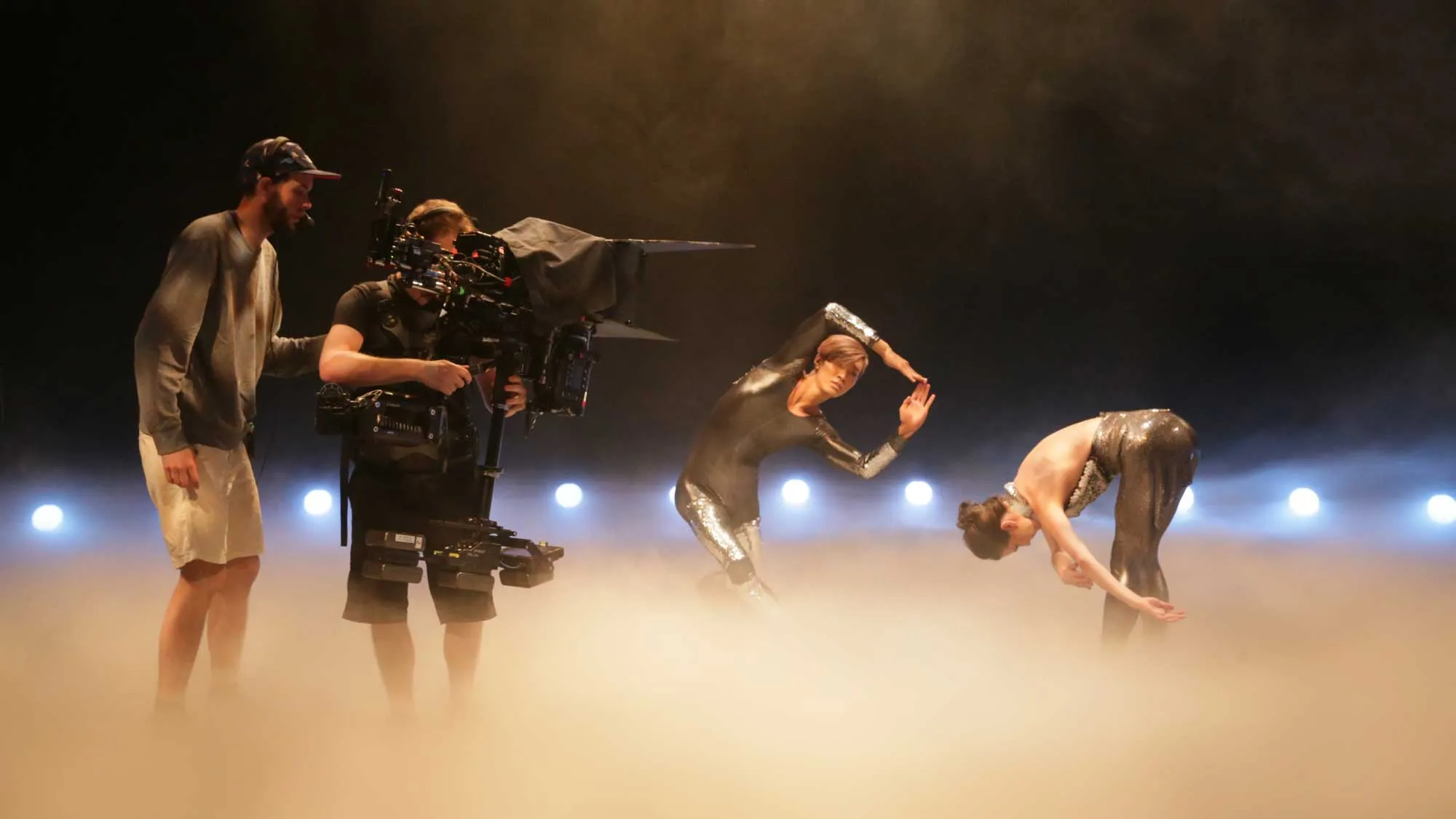 Two camera operators shooting two dancers wearing silver unitards standing in a sea of theatrical fog.