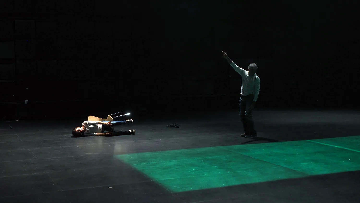 A man standing on a black stage pointing up as another person lays on the floor tipped over in their chair. A green rectangle is projected on to the floor.