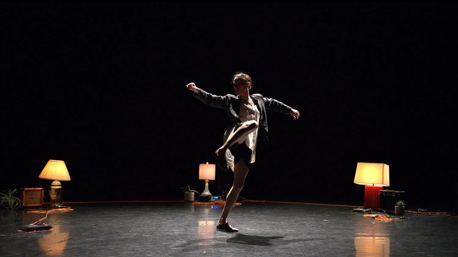 A female dancer wearing a white dress and blazer dancing with arms outstretched in a ring of orange extension cords and warmly lit lamps.
