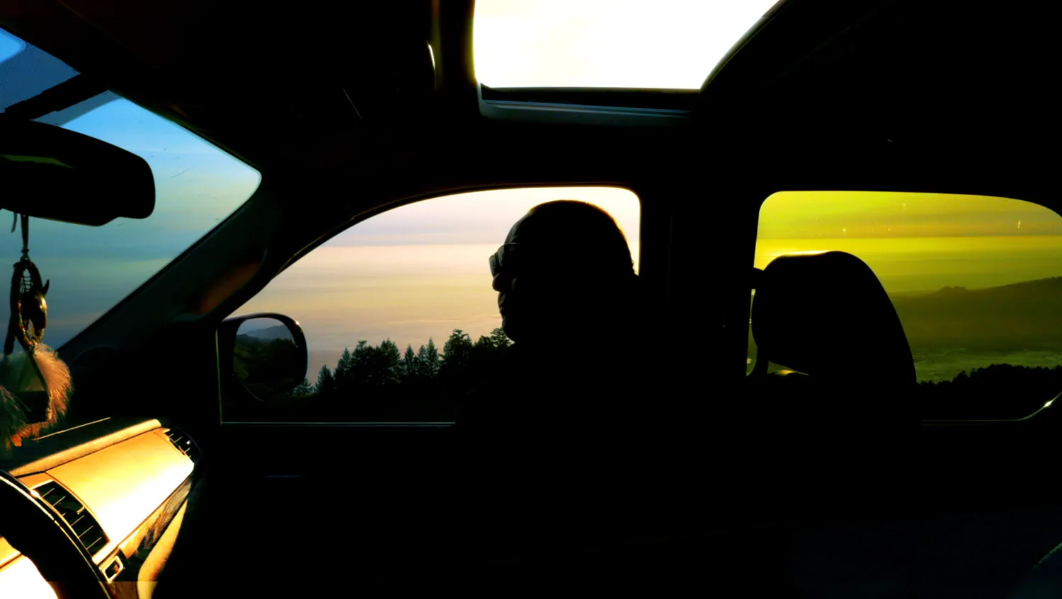 A person silhouetted in the passenger side of a car with the window and sunroof open 