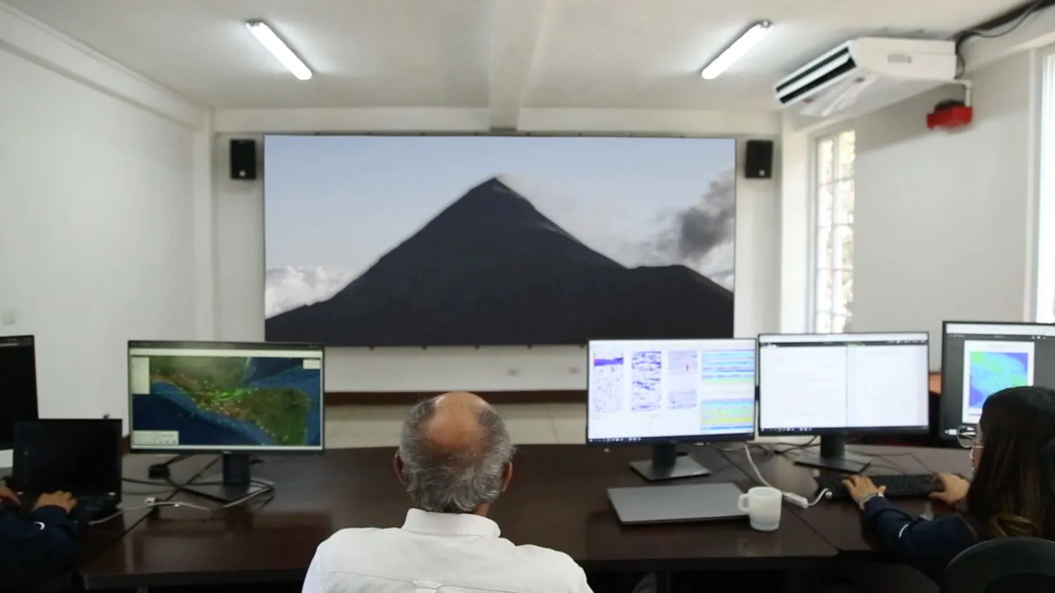 a man sitting in front of several computer screens looks at a large projection of a volcano