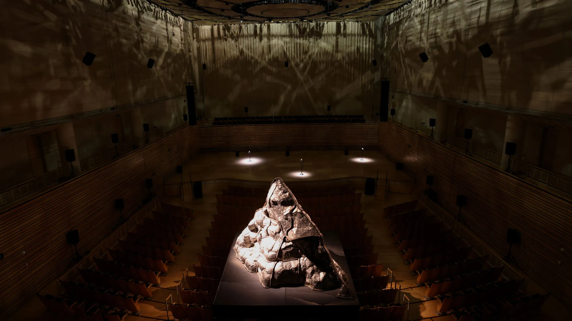 a steel volcano sits on a black platform in the middle of a concert hall with dramatic lighting from above