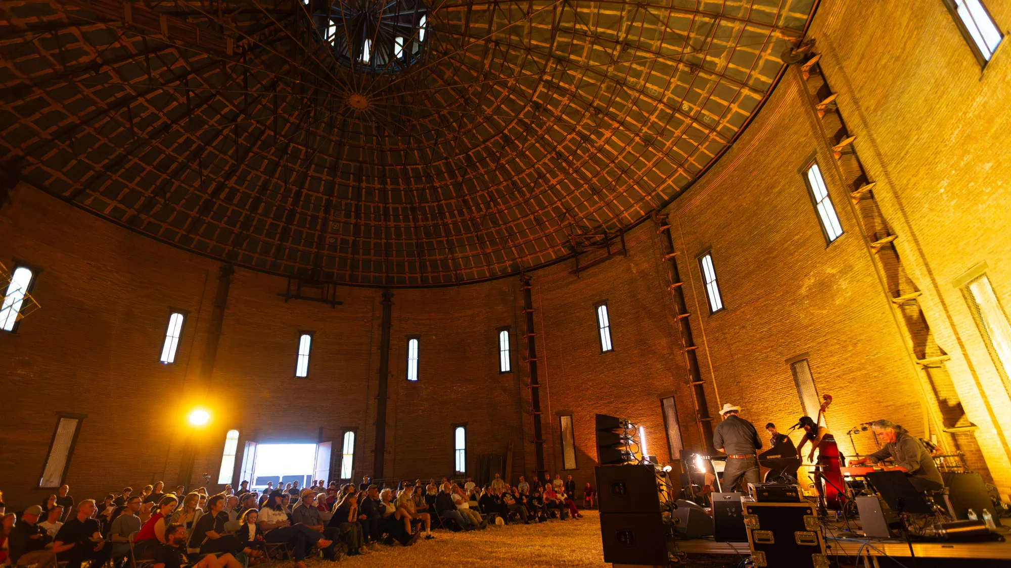 a crowd of concert-goers sits in chairs on hamster bedding in Troy's historic gasholder house while watching a five piece band.