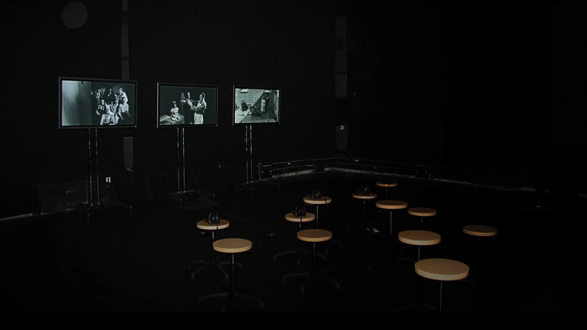 A dark black box theater with three small screens showing black and white images. Twelve round tan stools are are set up in front of them.