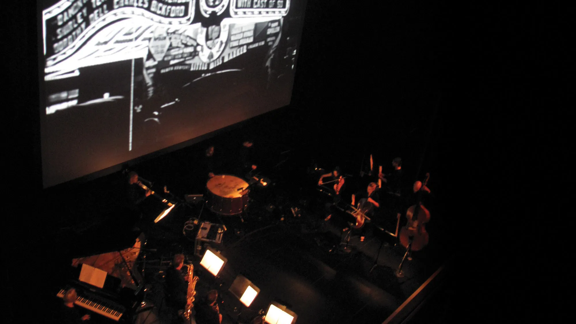 A pit orchestra in dim light playing at the foot of a large movie screen, showing a vintage black and white marquee.