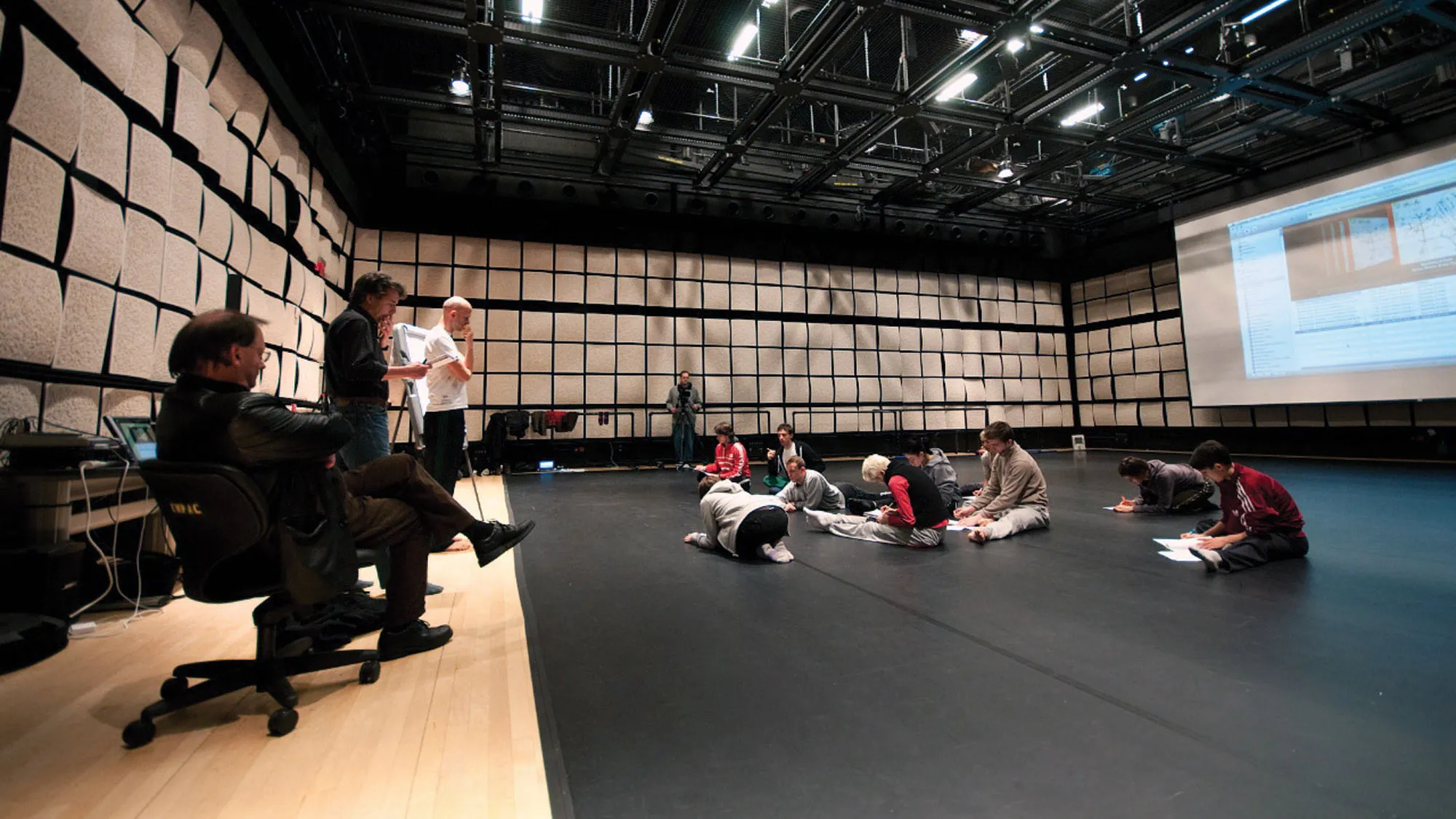 About ten dancers stretching on the floor of a studio as three men look on. 