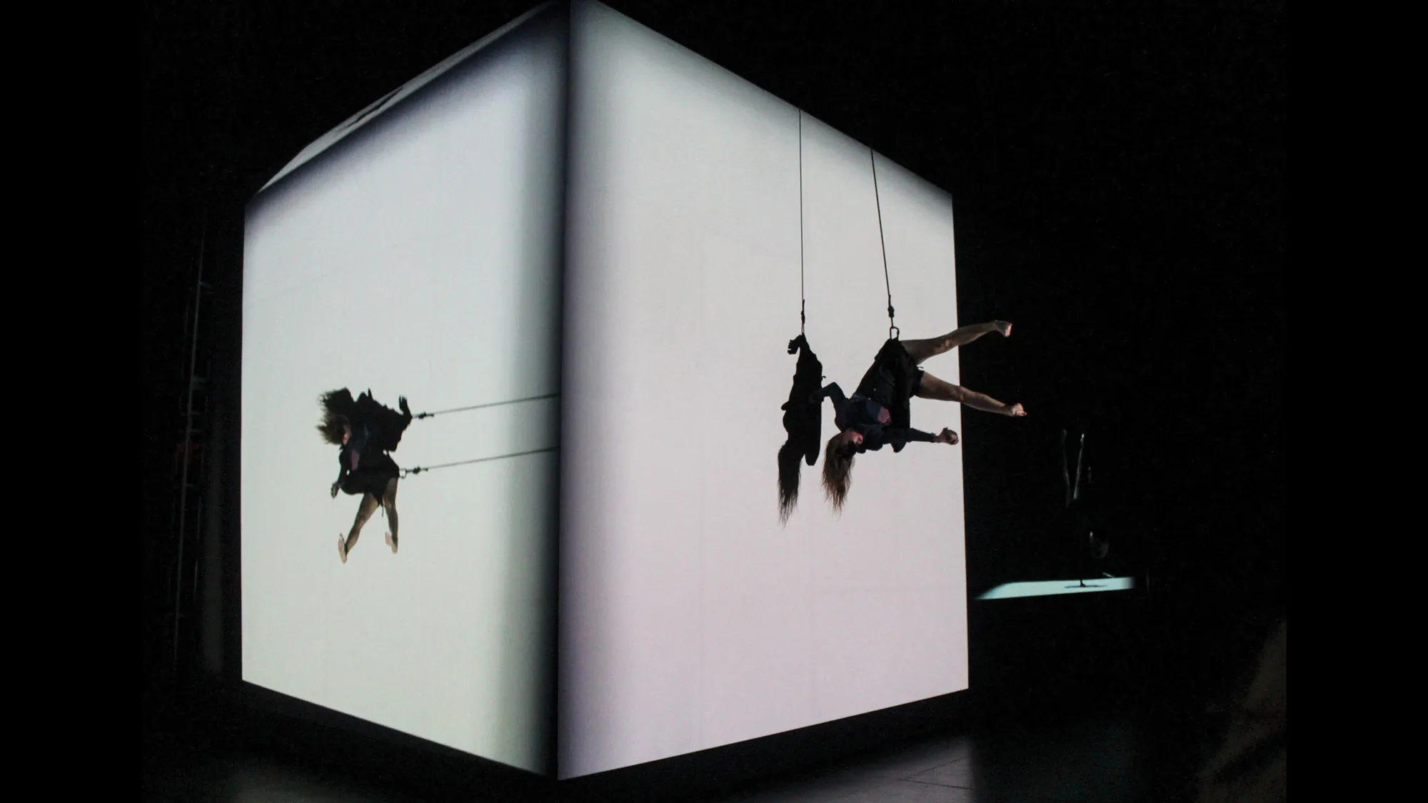 A woman suspended by a cable harness seemingly defying gravity while dancing on a white cube.