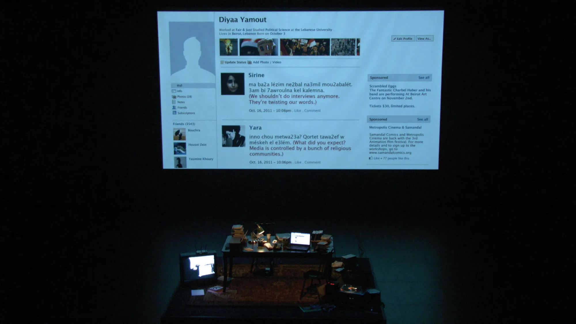 A cluttered desk on a black stage in front of a large screen projecting an image of an old Facebook profile. 