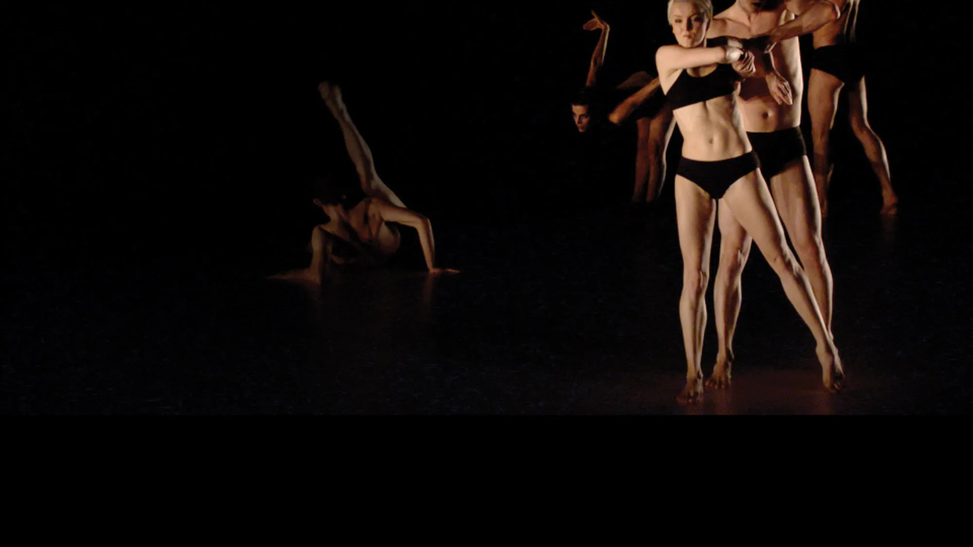 Five dancers wearing black underwear preforming on a black stage. A male dancer can be seen on the ground, back to the viewer with leg up as the rest are in a diagonal line.