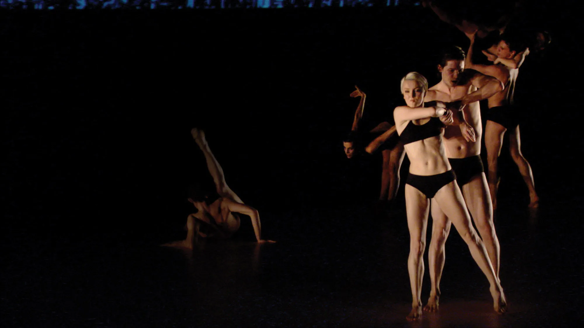 Six white dancers on dark stage dressed only in black underwear. Each person is in there own unique pose, including an individual on the floor with their leg extended and a woman standing in the front with arms twisted.