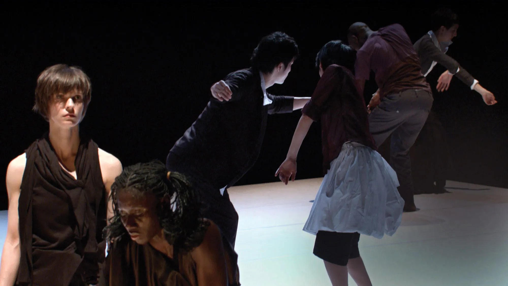Six dancers wearing street clothes moving throughout the space diagonally. A white woman and a Black woman stand in the foreground wearing black draped dresses. The white woman Is looking off into the distance as the Black woman hunches, looking down. 