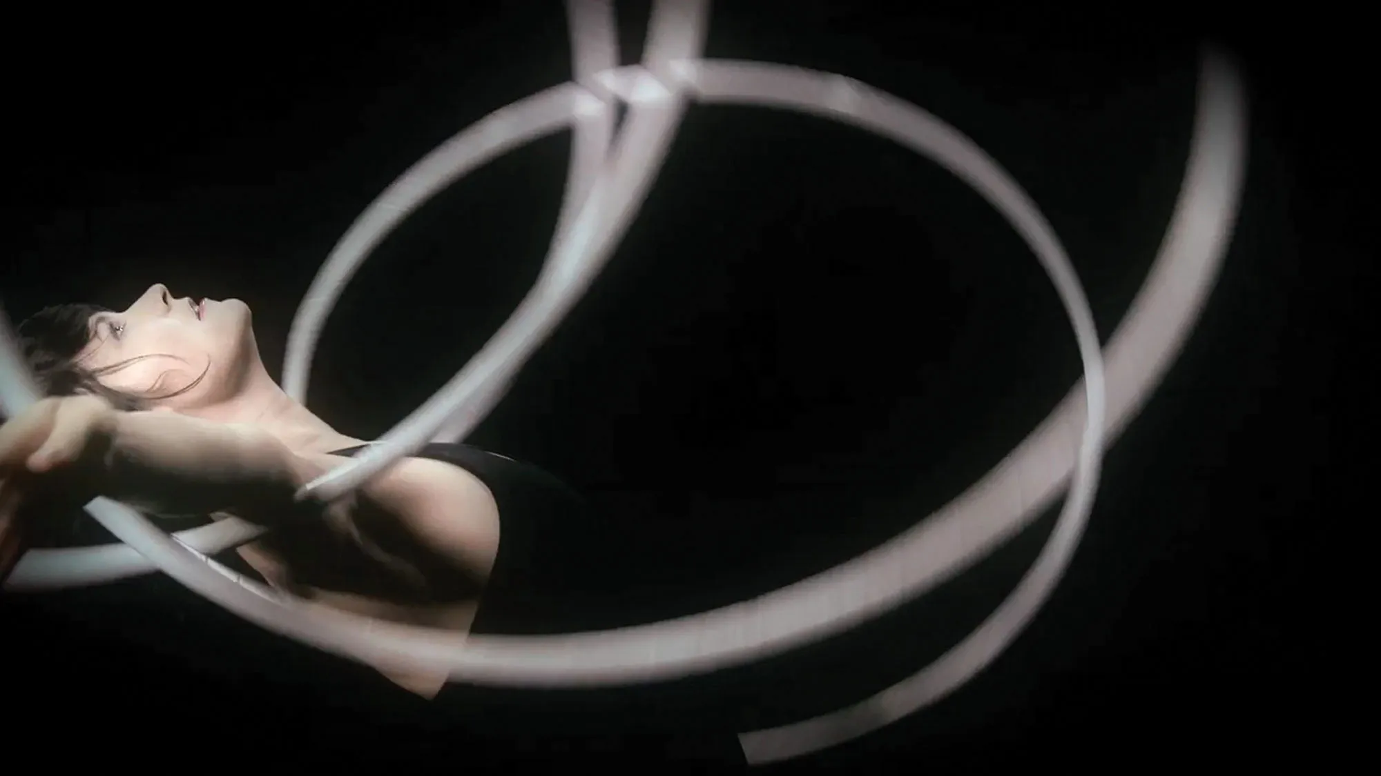 A white woman with dark hair arching her back and looking up with multiple white hoops spinning on her outstretched arms against a black background. 