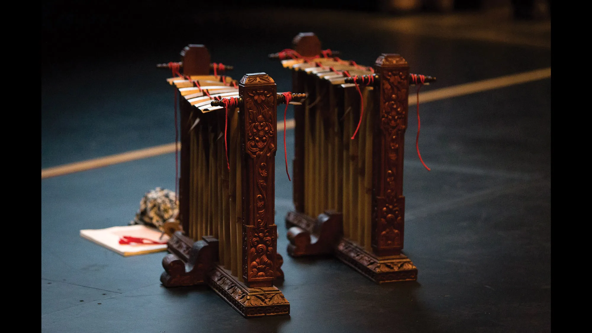 Two balinese instruments made of ornately carved wood held together by red string standing on a black floor. A small bouquet of flowers sits in front of them 