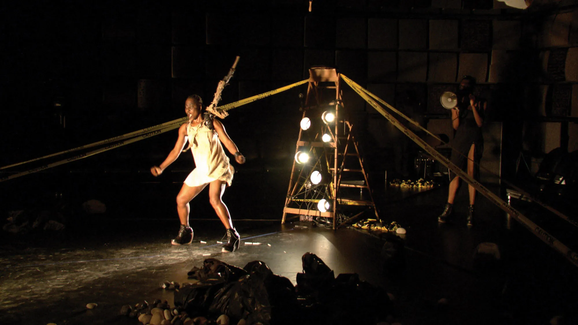 A Zimbabwean woman on a harness dancing in black box studio amongst piles of trash bags, caution tape, and a ladder with stage lights placed on the steps.