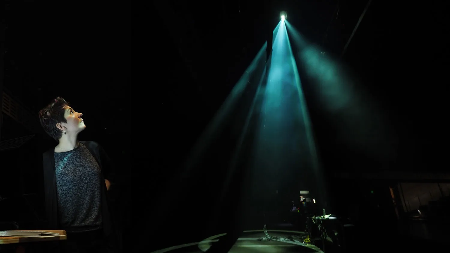Nina Young looking up dramatically at a blue beam of light o a dark stage.