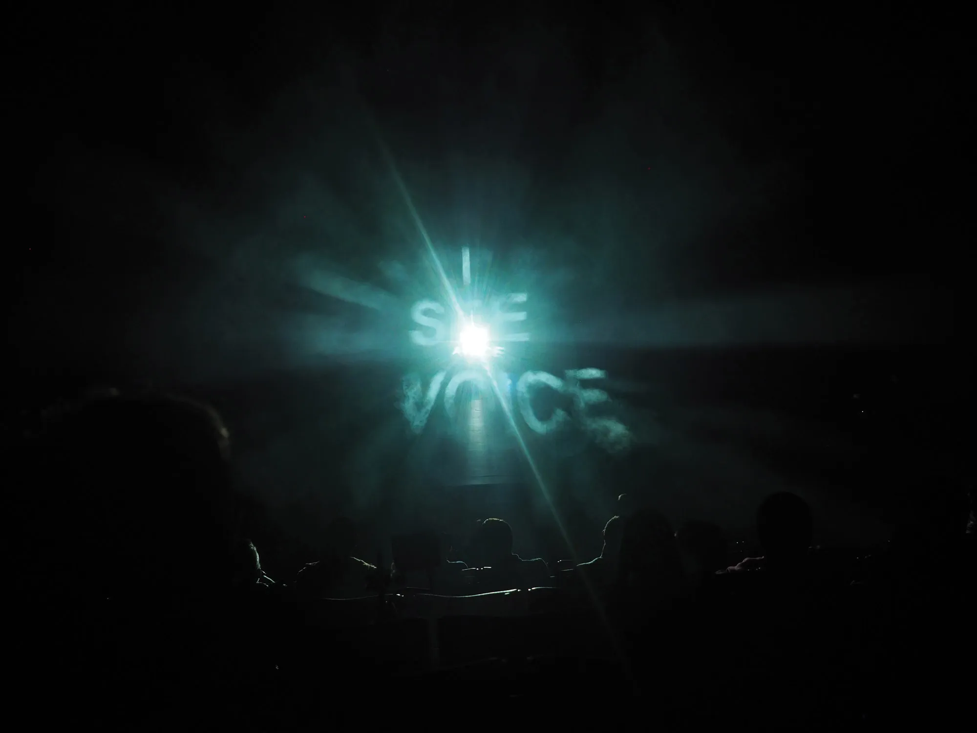 ghostly theatrical fog illuminates projected words "i see voice" while a silhouetted crowd seated in the theater looks on. 