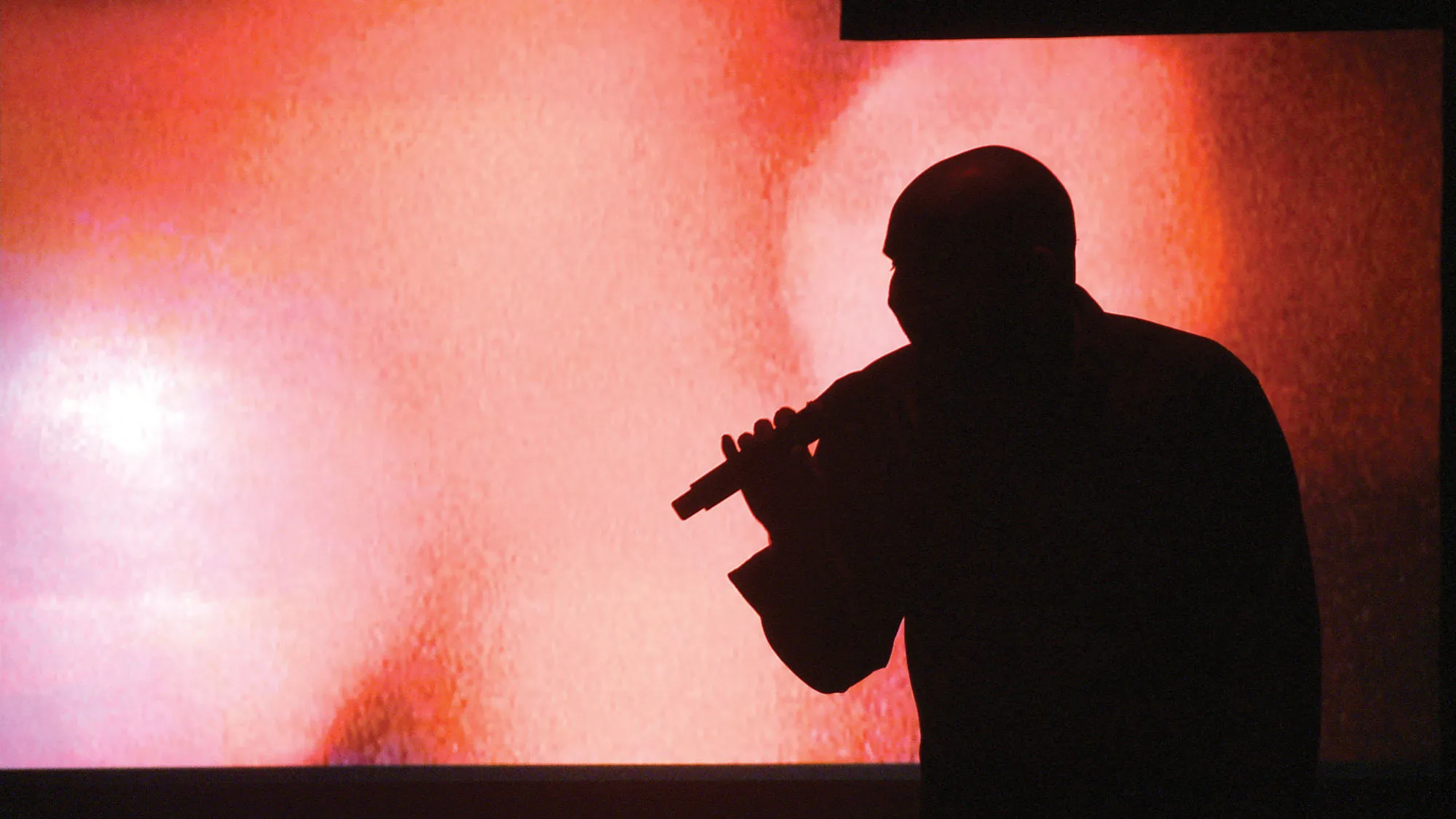 A bald person silhouetted against a red watercolored background holding a microphone. 