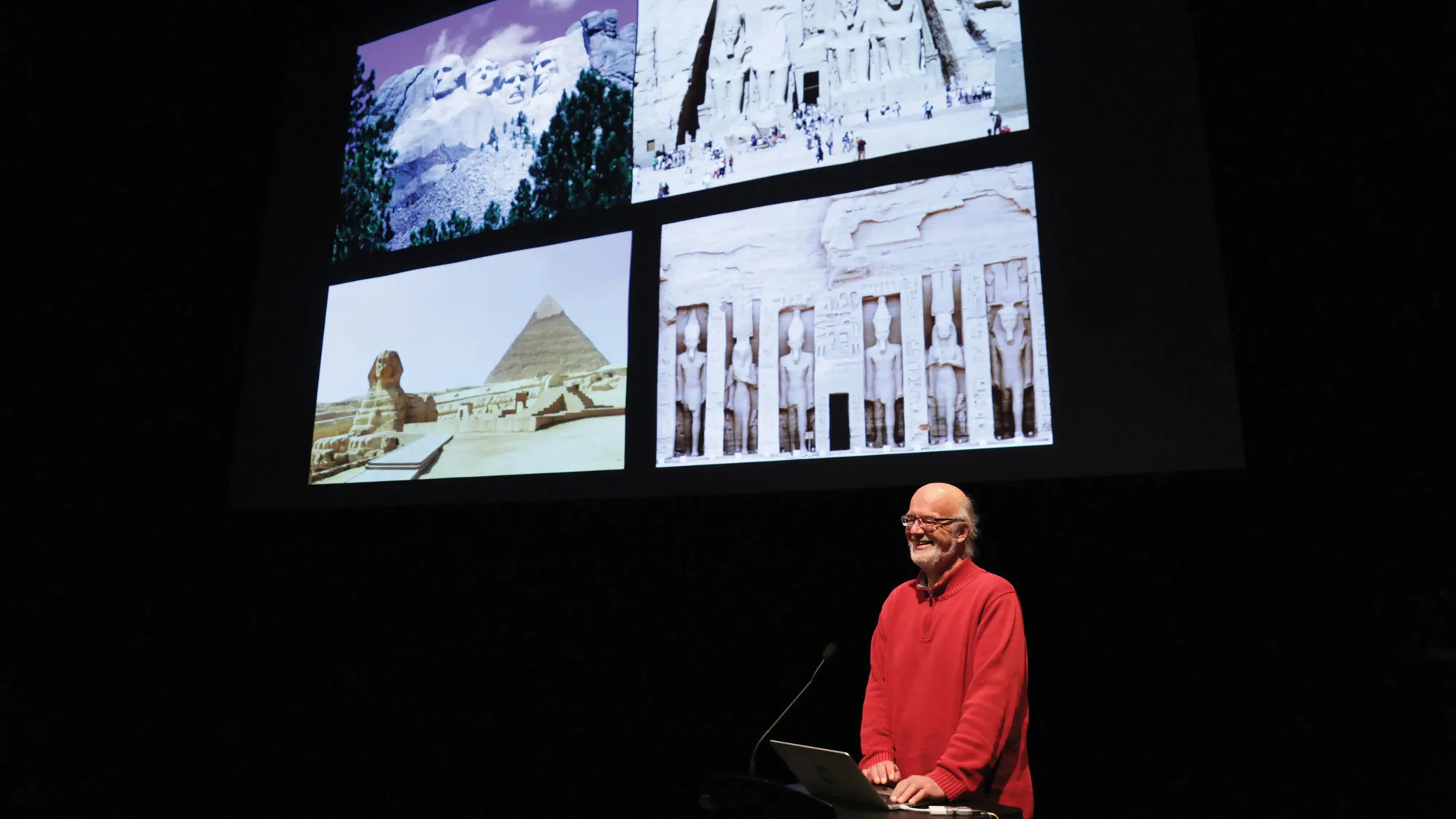 Johannes Goebel giving his talk on the Theater stage in front of a large screen displaying images of Mount Rushmore, The Sphinx, and other Egyptian monuments. 