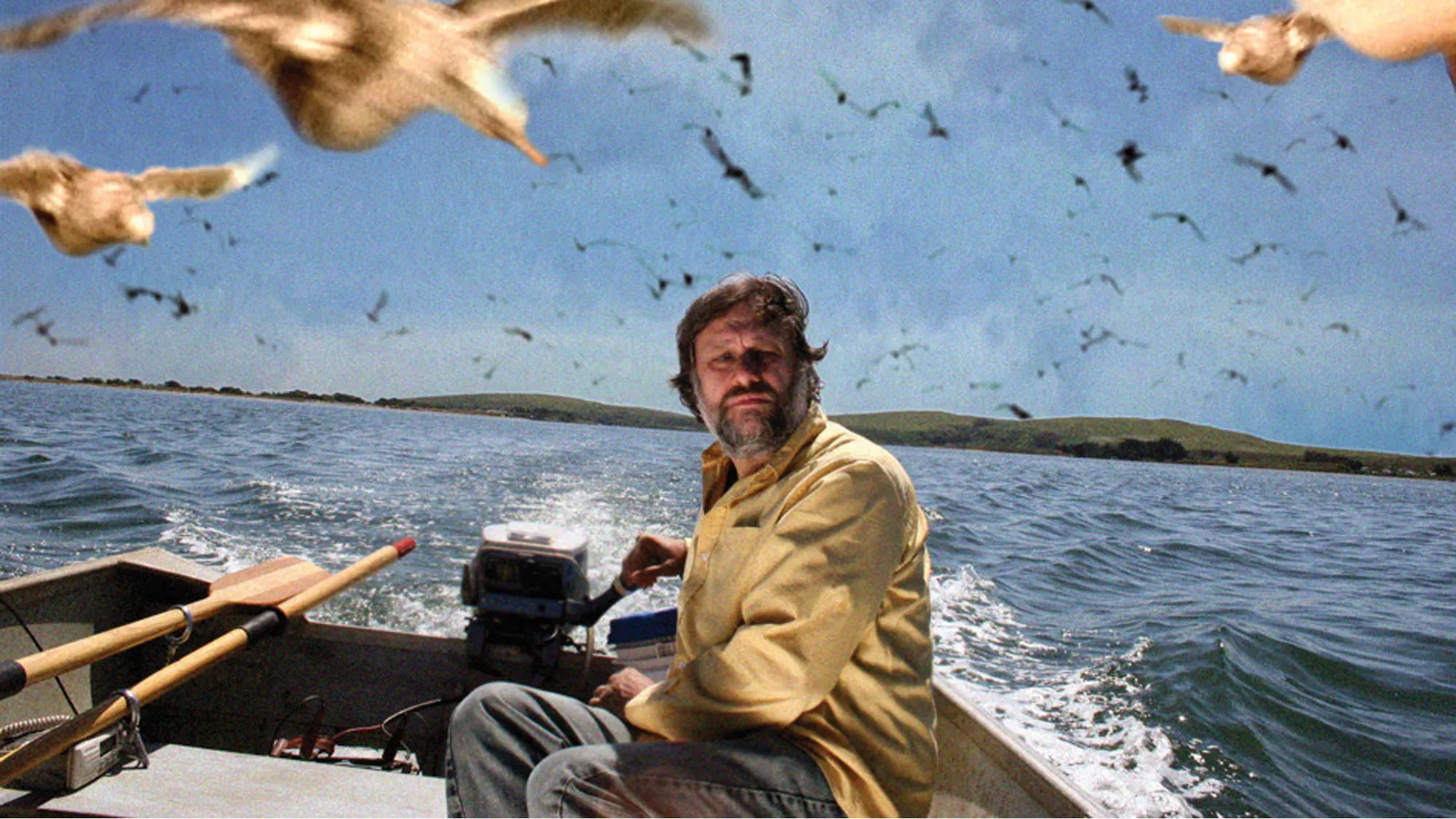 A white man with a greying beard and wearing a khaki coat and jeans, sitting at the back of a small fishing boat on the ocean surrounded by swooping gulls.