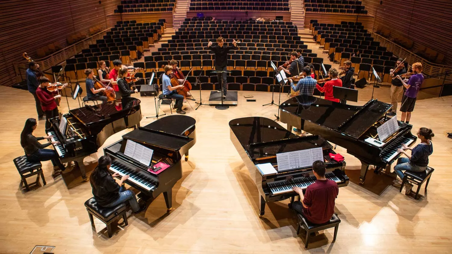 Four grand pianos on the concert hall stage along with a small string orchestra and conductor in rehearsal. 