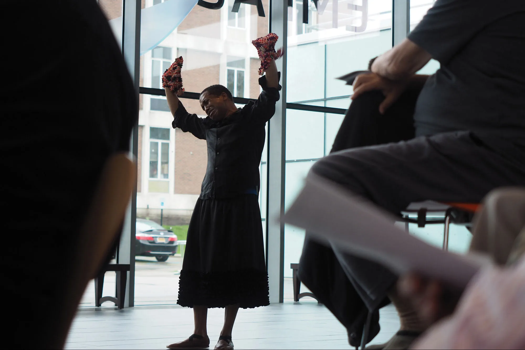 Trajal Harrell wearing a black dress performing with hands up in a light filled lobby. 
