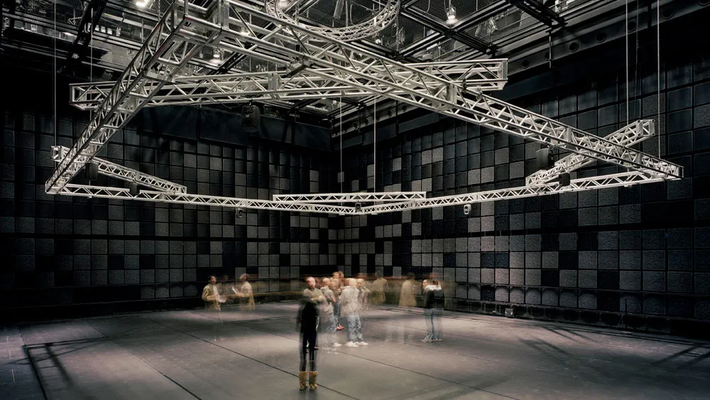 A crowd blurred in motion under rigging suspended from the ceiling of a black box theater.